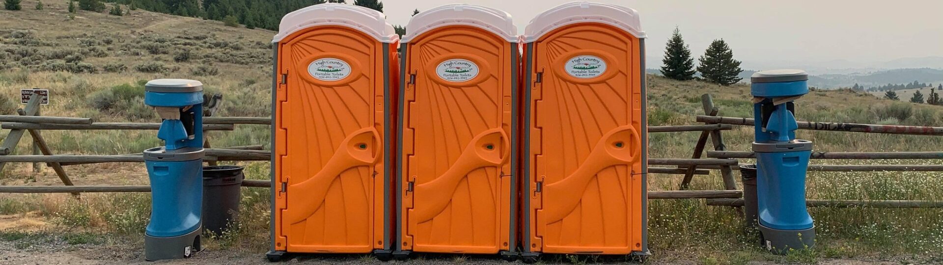 Clean orange portable toilets and blue wash stations lined up along a ranch fence with the Montana high country behind